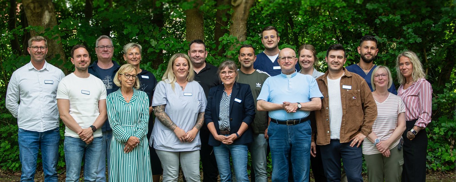 Ein Gruppenfoto von etwa 17 Personen, die lächelnd im Freien vor einem bewaldeten Hintergrund stehen. Die Gruppe besteht aus Männern und Frauen unterschiedlichen Alters in lässiger bis geschäftlicher Freizeitkleidung.