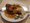 Ein weißer Teller auf einem Holztisch zeigt ein Hauptgericht mit gebratenem Fleisch, Reis und gemischtem Gemüse. Im unscharfen Hintergrund sitzt eine weitere Person mit einem ähnlichen Essen.