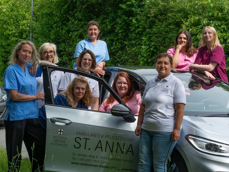 Ein Team von neun Frauen im medizinischen Bereich posiert lächelnd um einen silbernen Dienstwagen. Auf der Tür steht „Ambulanter Pflegedienst St. Anna“. Die Frauen tragen blaue, rosa und weiße Arbeitskleidung.