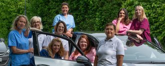 Ein Team von neun Frauen im medizinischen Bereich posiert lächelnd um einen silbernen Dienstwagen. Auf der Tür steht „Ambulanter Pflegedienst St. Anna“. Die Frauen tragen blaue, rosa und weiße Arbeitskleidung.
