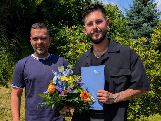 Zwei Männer stehen im Freien vor grüner Vegetation. Der Mann links hält einen bunten Blumenstrauß, der Mann rechts eine blaue Mappe. Beide lächeln in die Kamera.