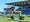 Ein Reiter auf einem braunen Pferd springt über ein grünes Hindernis in einem Stadion. Im Hintergrund sind Tribünen und Flaggen unter blauem Himmel zu sehen.