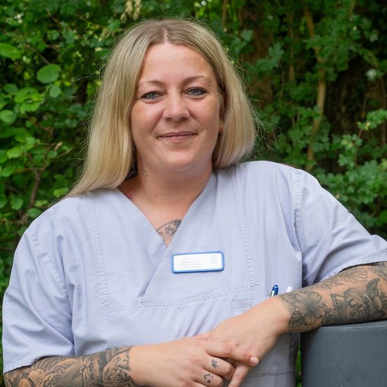 Porträt einer lächelnden Frau mit blonden Haaren und sichtbaren Tattoos an den Armen. Sie trägt eine hellblaue medizinische Arbeitskleidung und lehnt vor einem grünen, natürlichen Hintergrund.