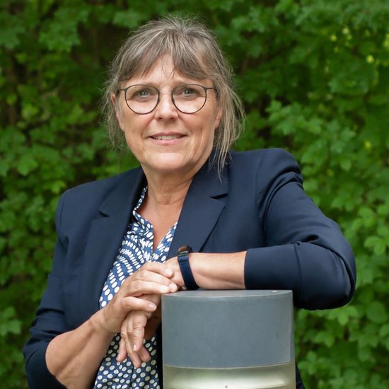 Eine Frau mit Brille und dunklem Blazer lächelt in die Kamera. Sie lehnt mit verschränkten Armen auf einer zylindrischen Gartenlampe vor einem grünen, unscharfen Hintergrund.