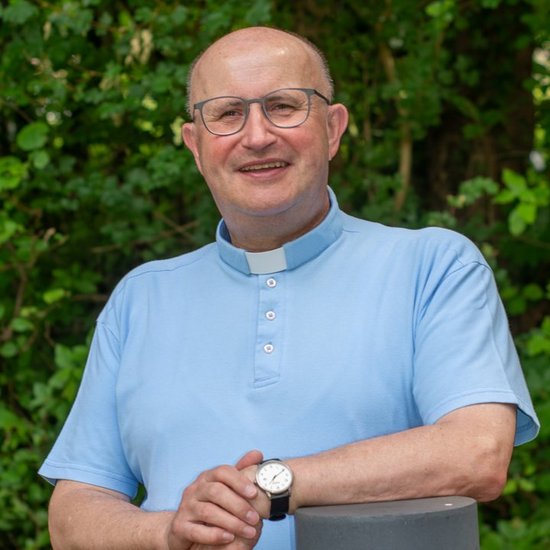 Ein lächelnder Priester mit Brille und hellblauem Kragenhemd lehnt sich lässig an einen grauen Pfosten vor einem grünen, bewachsenen Hintergrund.