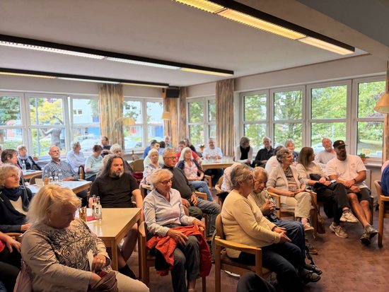 Eine Gruppe von Menschen, überwiegend ältere Erwachsene, sitzt in einem hellen Raum mit vielen Fenstern und scheint einer Präsentation oder einem Vortrag beizuwohnen.