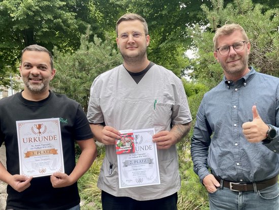 Drei Männer stehen im Freien vor Bäumen. Zwei von ihnen halten Urkunden in die Kamera, die wie Auszeichnungen wirken. Der Mann in der Mitte trägt graue Kleidung. Der Mann rechts zeigt ein Daumen-hoch-Zeichen.