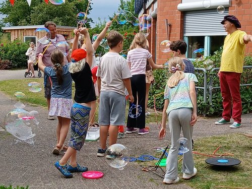 Kinder und Erwachsene spielen im Freien mit riesigen Seifenblasen. Sie stehen auf einem gepflasterten Hof vor einem Backsteingebäude und strecken die Arme aus, um die schwebenden Blasen zu erreichen. Rote und weiße Wimpelketten hängen über dem Platz.