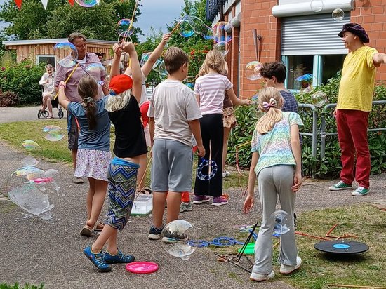 Kinder und Erwachsene spielen im Freien mit riesigen Seifenblasen. Sie stehen auf einem gepflasterten Hof vor einem Backsteingebäude und strecken die Arme aus, um die schwebenden Blasen zu erreichen. Rote und weiße Wimpelketten hängen über dem Platz.