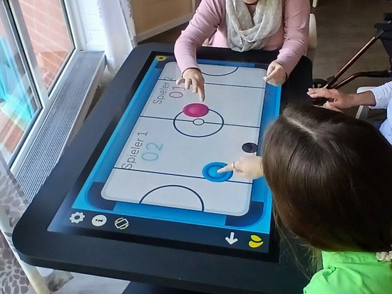 Eine ältere Frau in einem rosa Cardigan und ein Kind in einem grünen Shirt spielen gemeinsam ein digitales Tischspiel auf einem großen Touchscreen-Tisch. Sie sitzen sich gegenüber und berühren den Bildschirm.